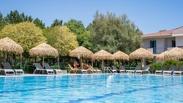 Ein Swimmingpool mit Liegestühlen und Stroh-Sonnenschirmen, umgeben von Bäumen und einem Gebäude mit Ziegeldach.