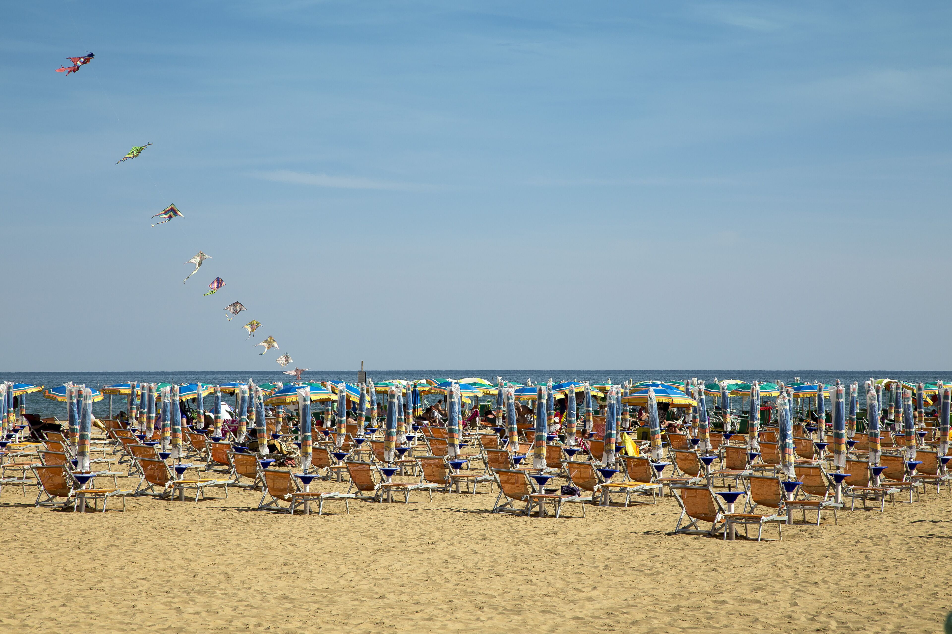 A beach with rows of sun loungers and umbrellas, and a clear sky with kites flying.