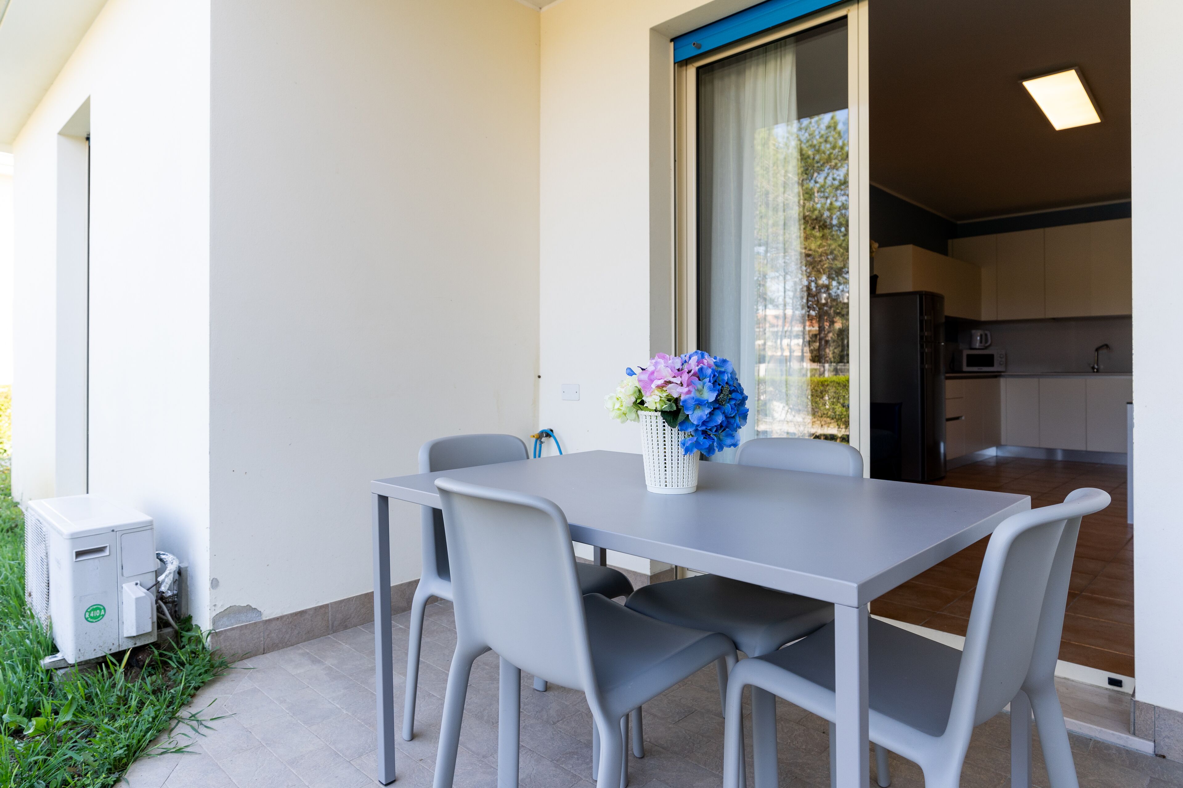 Ein moderner Essbereich mit einem grauen Tisch und Stühlen, einer weißen Vase mit Blumen und Blick auf eine Küche durch ein großes Fenster.