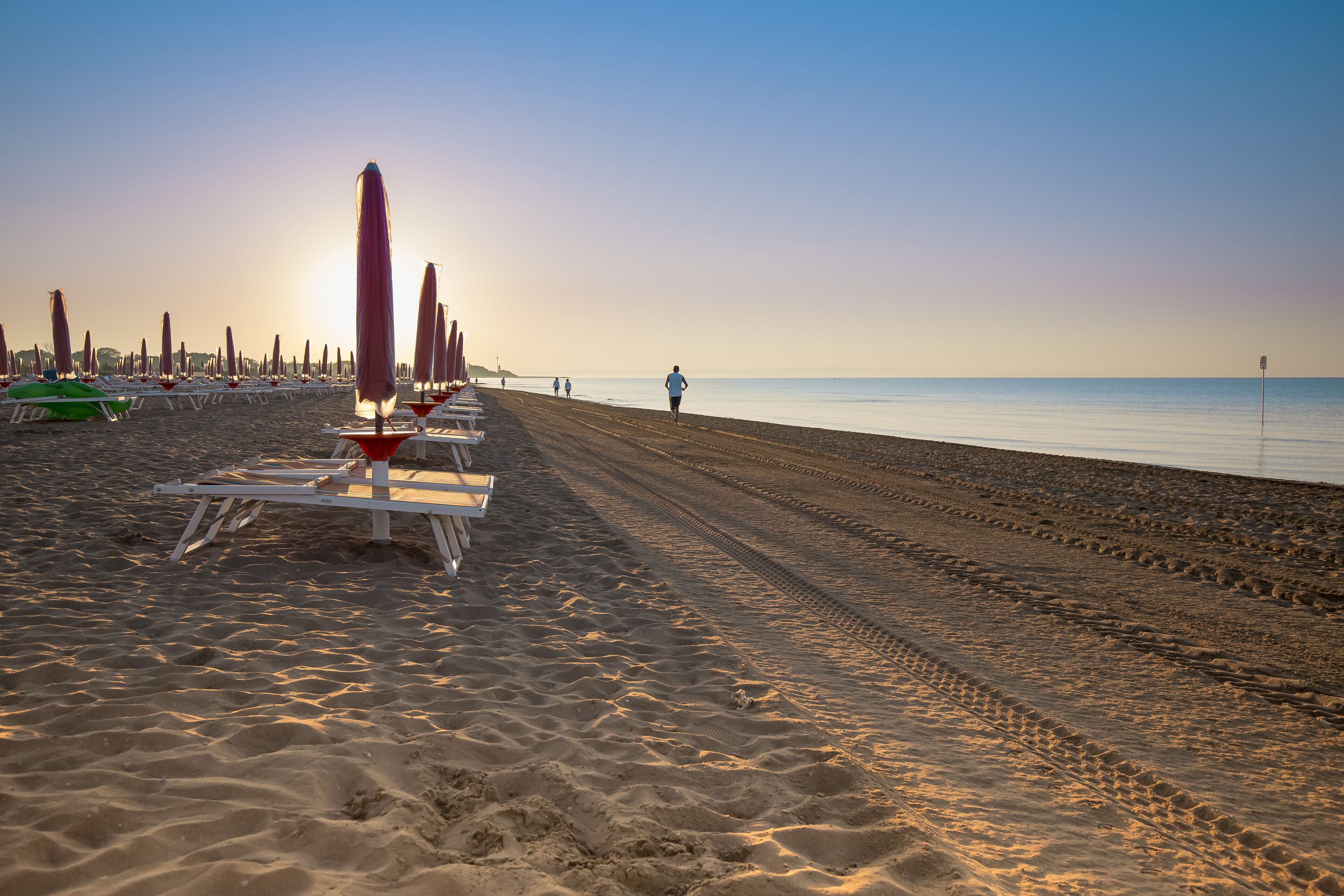 A beach with rows of sun loungers and umbrellas, a clear sky, and a person walking along the shore.