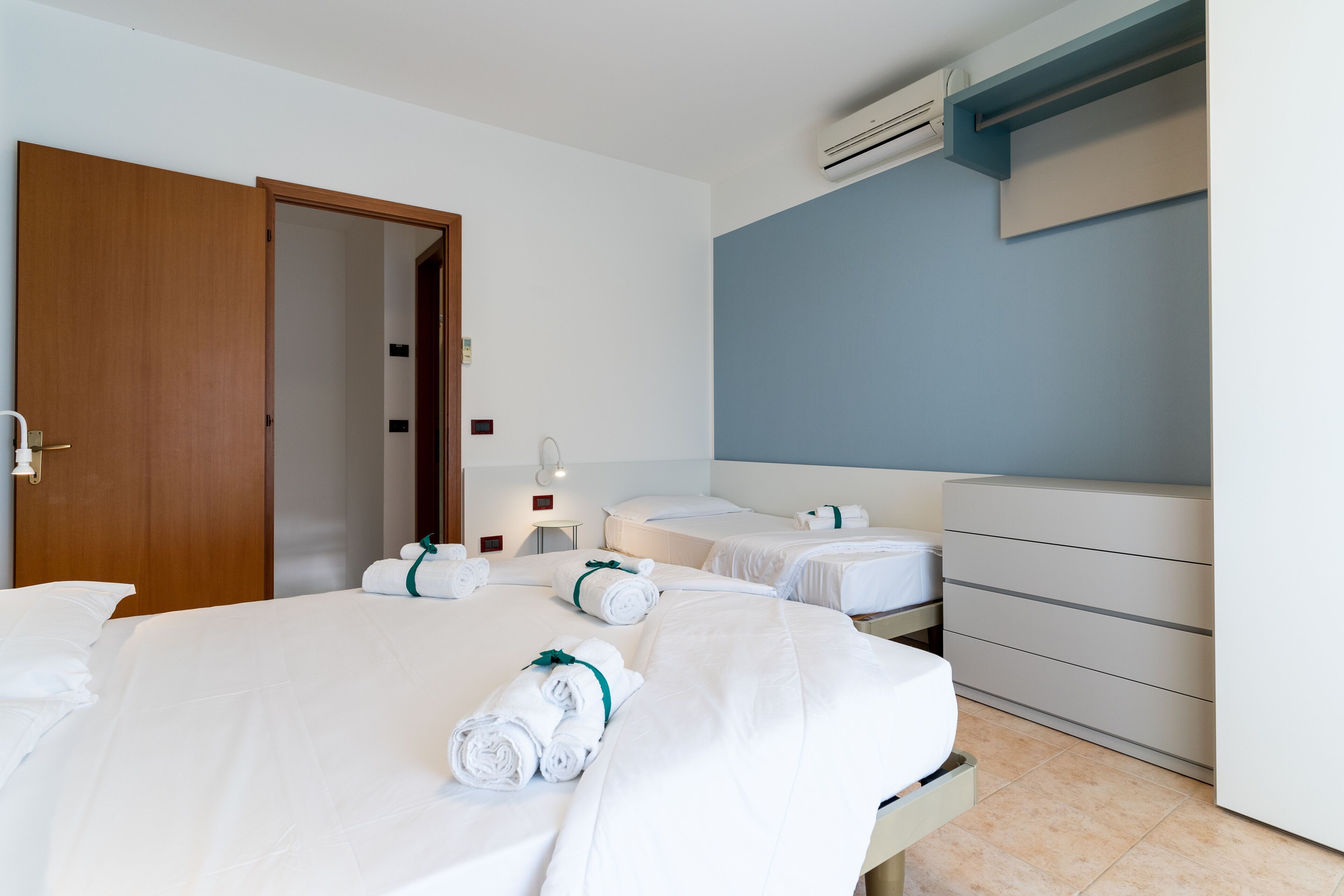 Una camera d'albergo con due letti, una porta in legno, un comò bianco e un condizionatore d'aria.