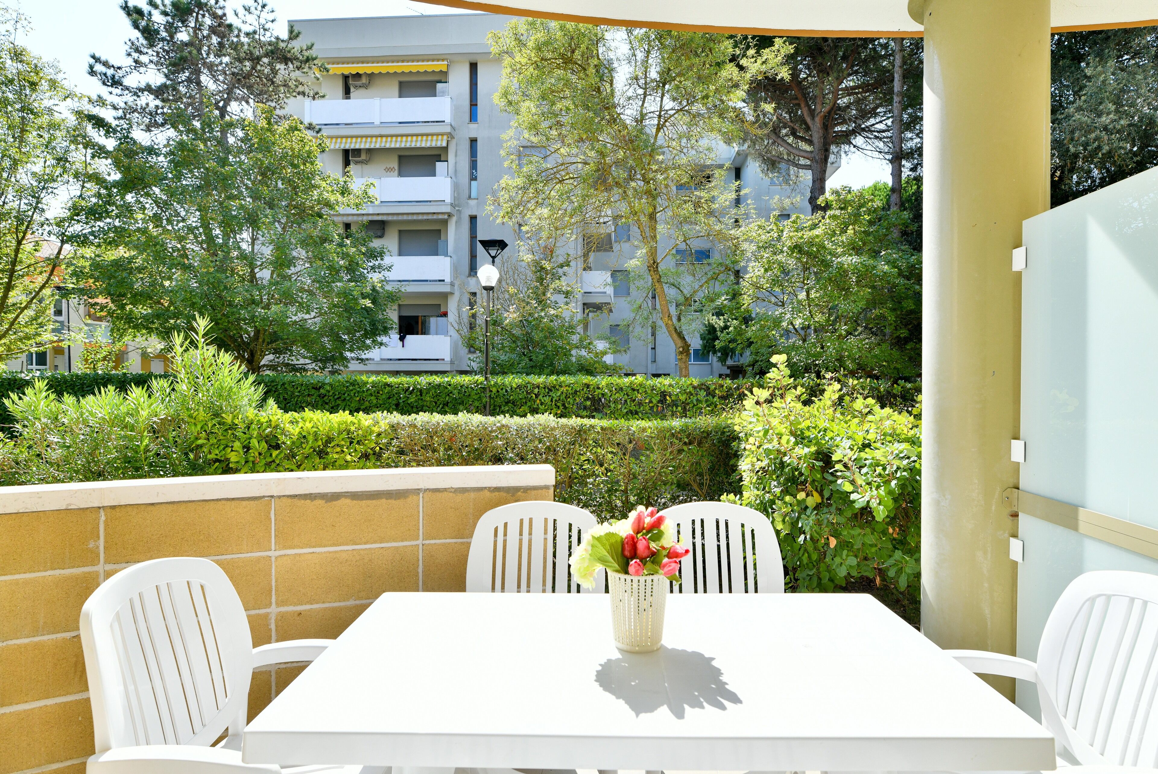 An outdoor patio with a white table and chairs, a vase with flowers, and a view of a multi-story building with balconies.