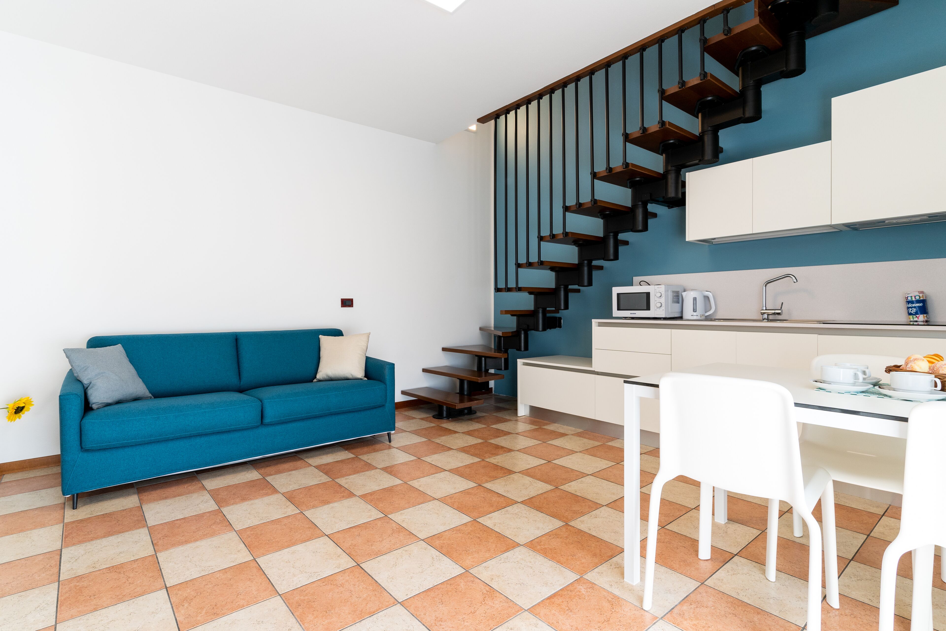 A modern kitchen with a staircase, a dining area with white chairs, and a blue sofa with a cushion.