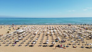 Ein Strand mit vielen Liegestühlen und Sonnenschirmen, einem klaren Himmel und dem Meer im Hintergrund.