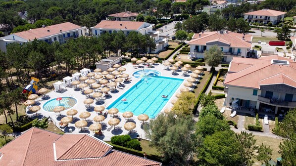 Um resort com uma grande piscina cercada por espreguiçadeiras e guarda-sóis, vários edifícios com telhados em vermelho e uma área de jardim bem cuidada.