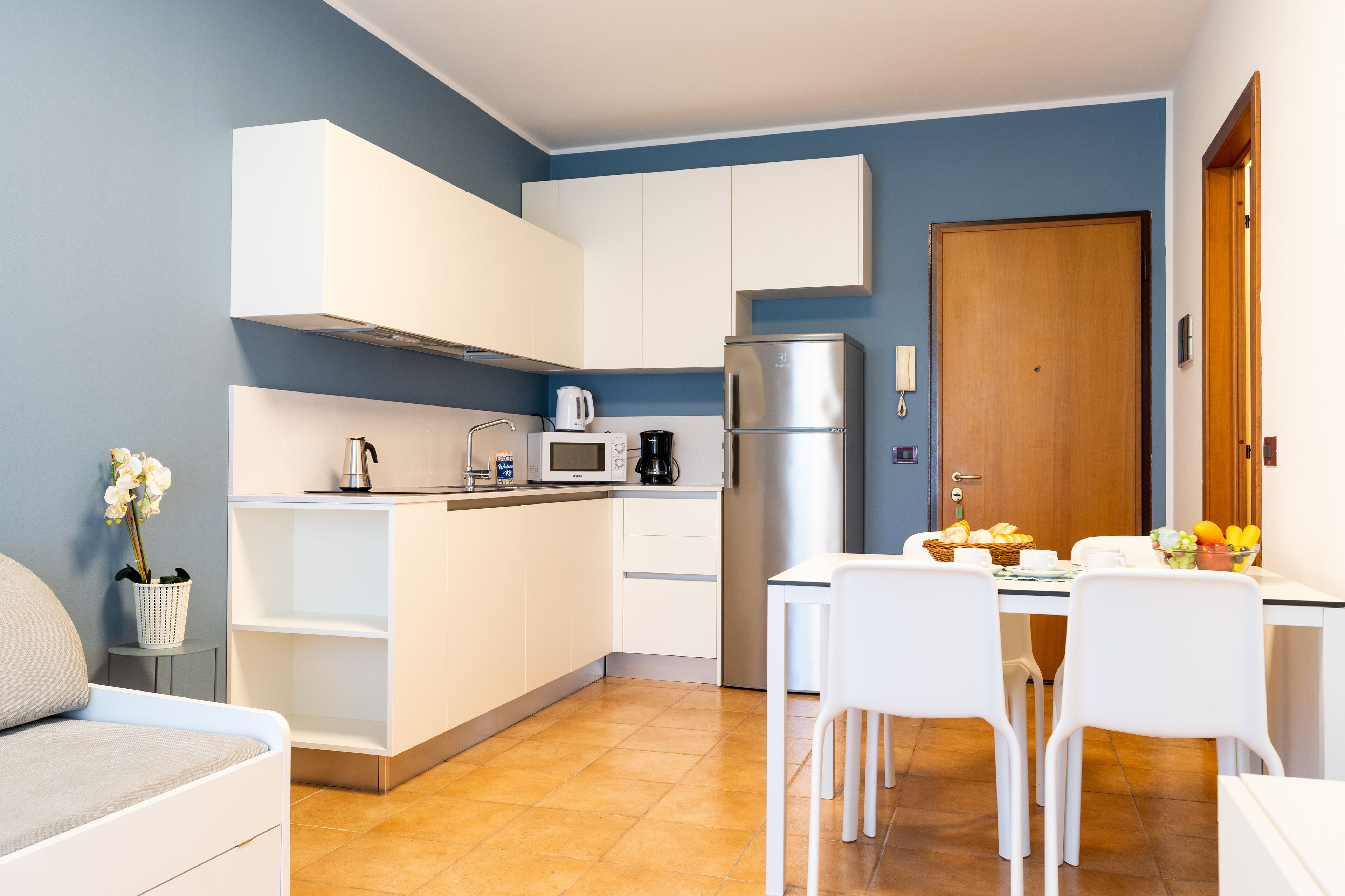 A compact kitchen with white cabinets, a stainless steel refrigerator, a microwave, and a coffee maker. There is a dining table with chairs and a basket of bread.