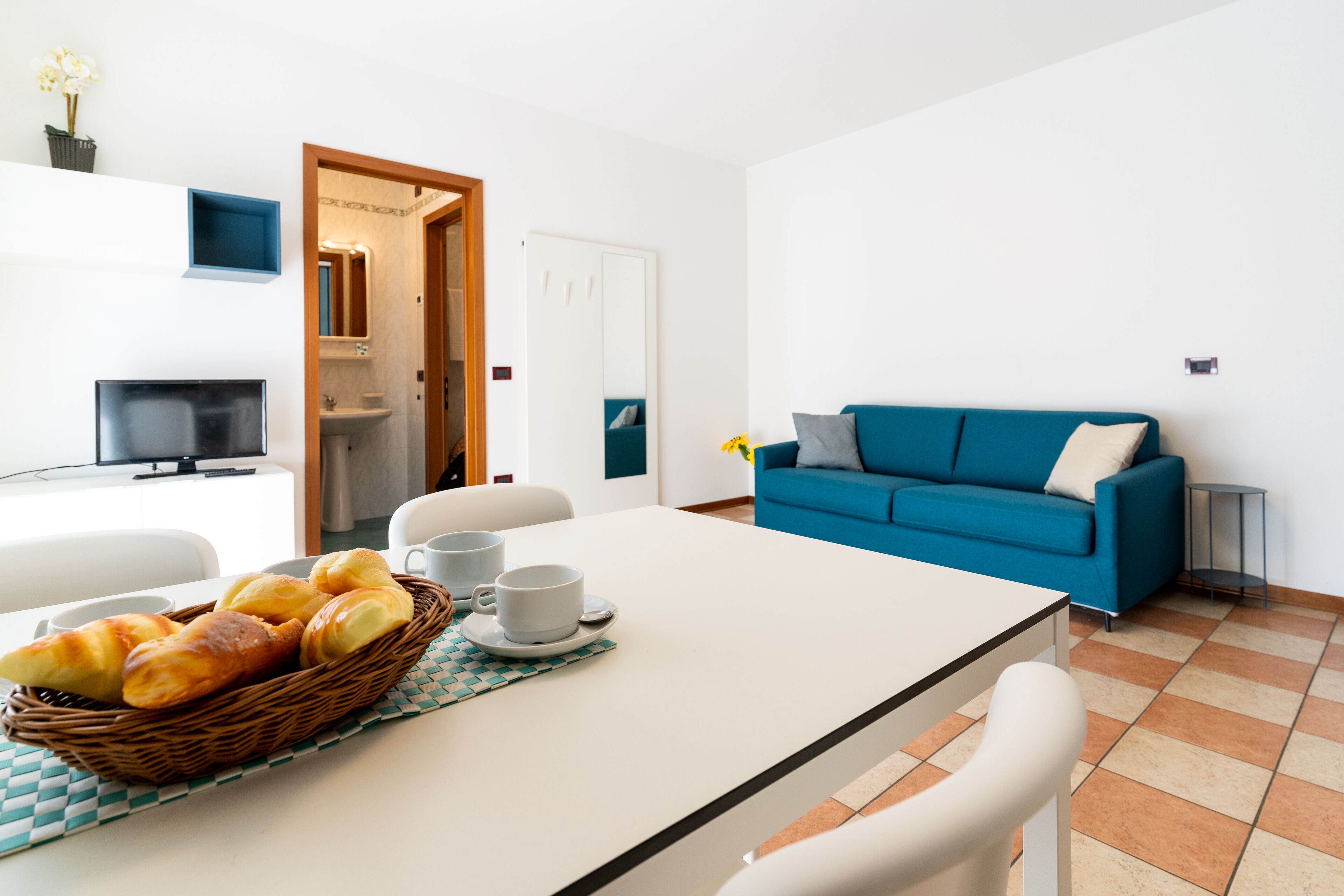 Una sala de estar moderna con un sofá azul, una mesa de comedor con pan y tazas, y un televisor en una esquina.