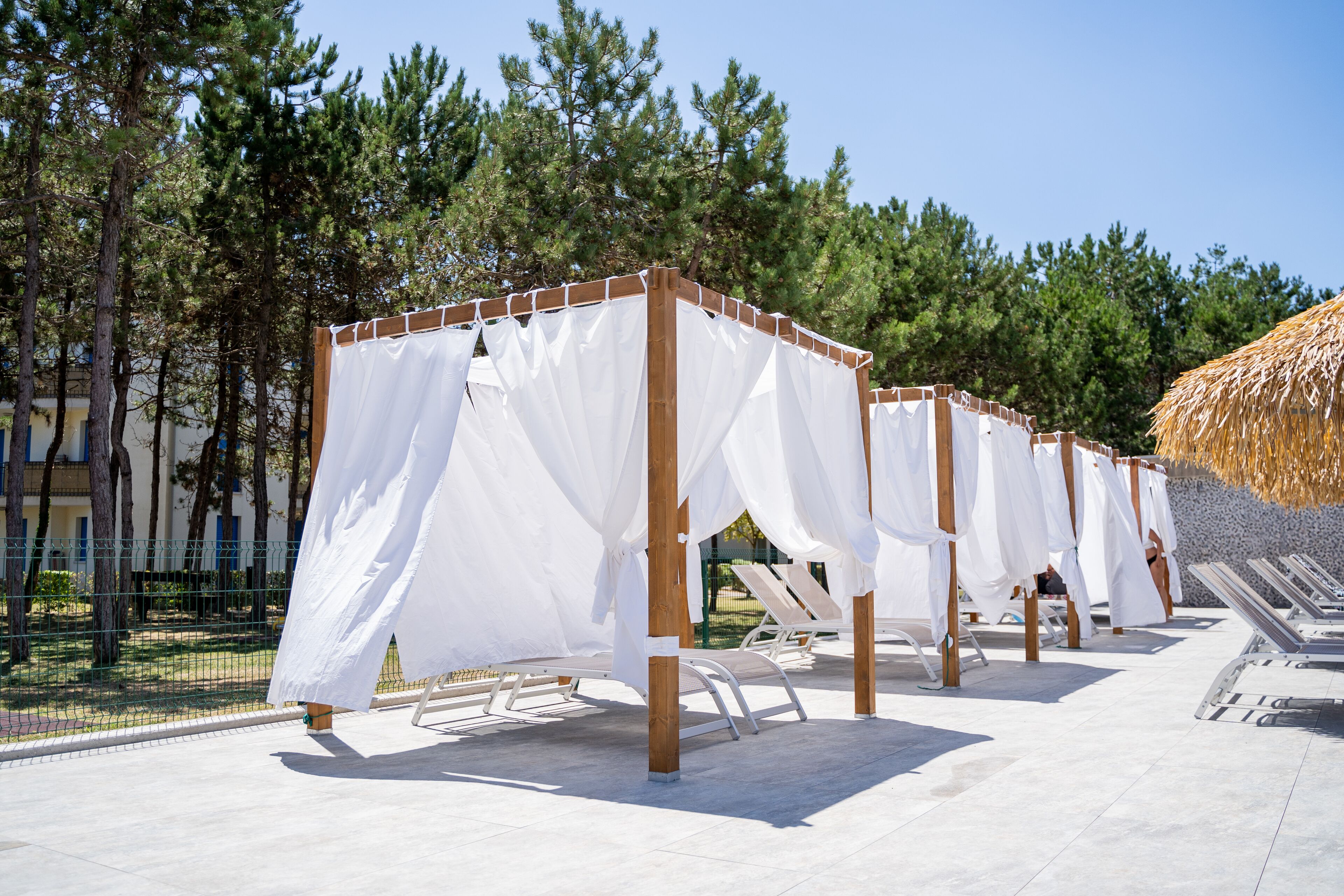 Une rangée de tentes blanches à toit tendu, dotées d’une structure en bois, est installée sur une terrasse carrelée, agrémentée de chaises longues.