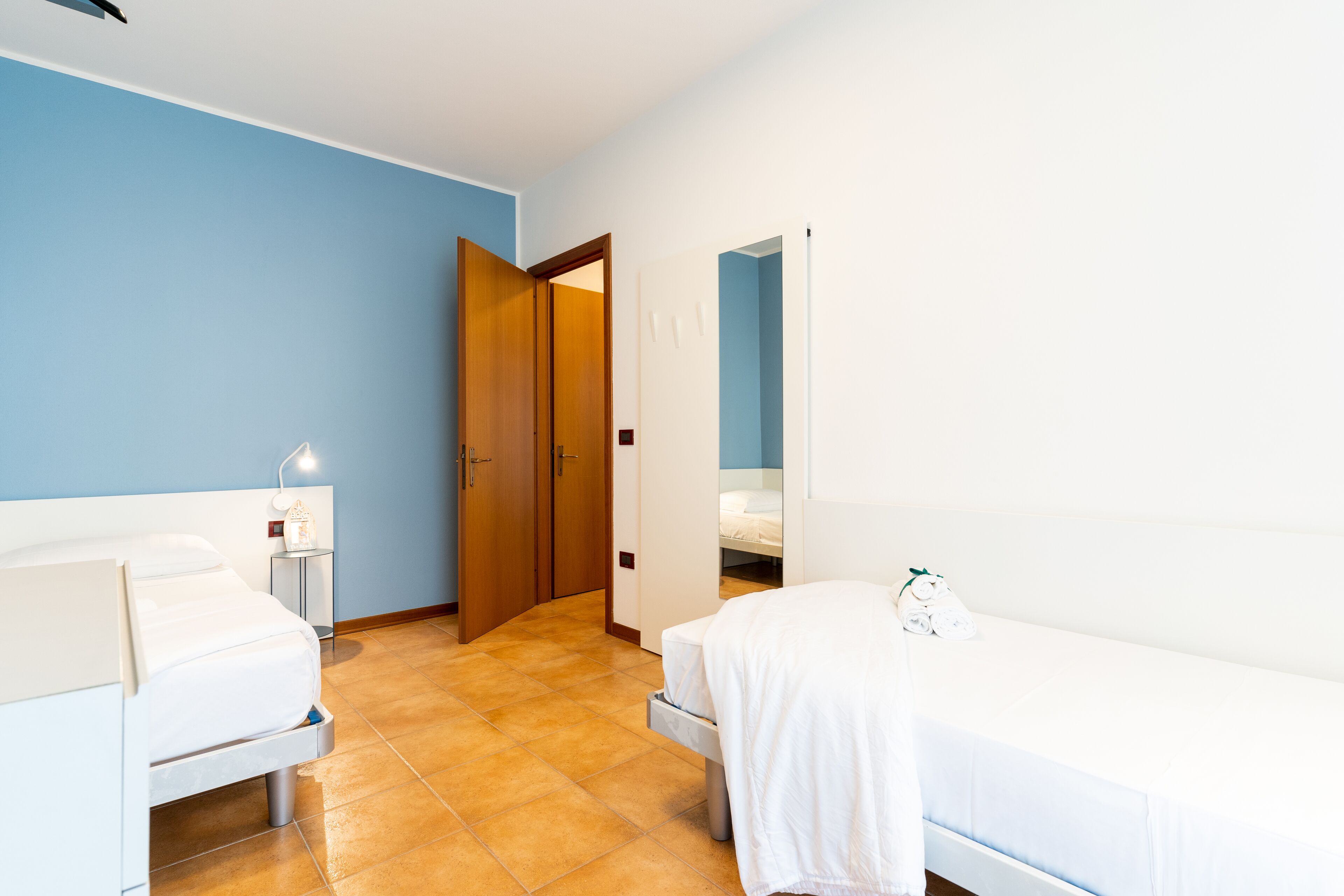 A hotel room with two beds, a bedside table with a lamp, and a door leading to another room.