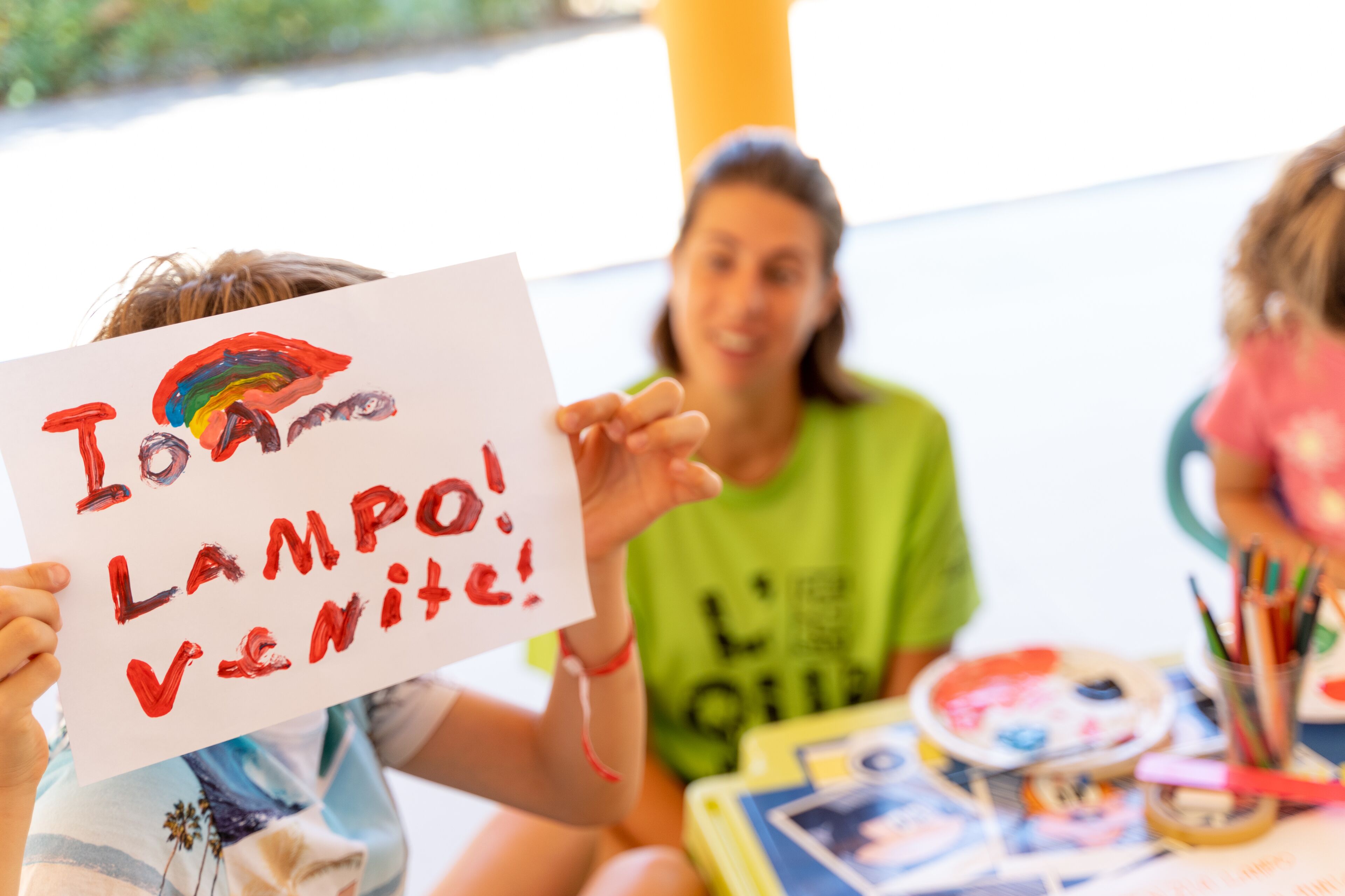 A child holding a sign with a rainbow and the words "I love LAMP! VENITE!" in a classroom setting with art supplies on the table.