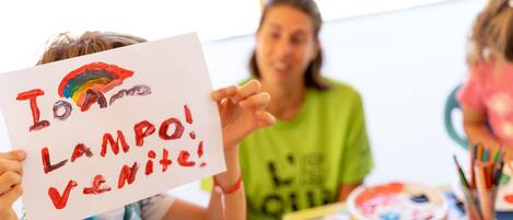 A child holding a sign with a rainbow and the words "I love LAMP! VENITE!" in a classroom setting with art supplies on the table.
