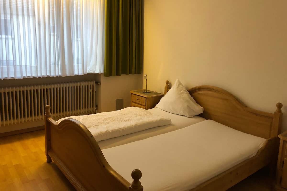 Un lit double avec une tête de lit en bois, une table de chevet avec une lampe, un radiateur et des rideaux légers avec un drapé vert.