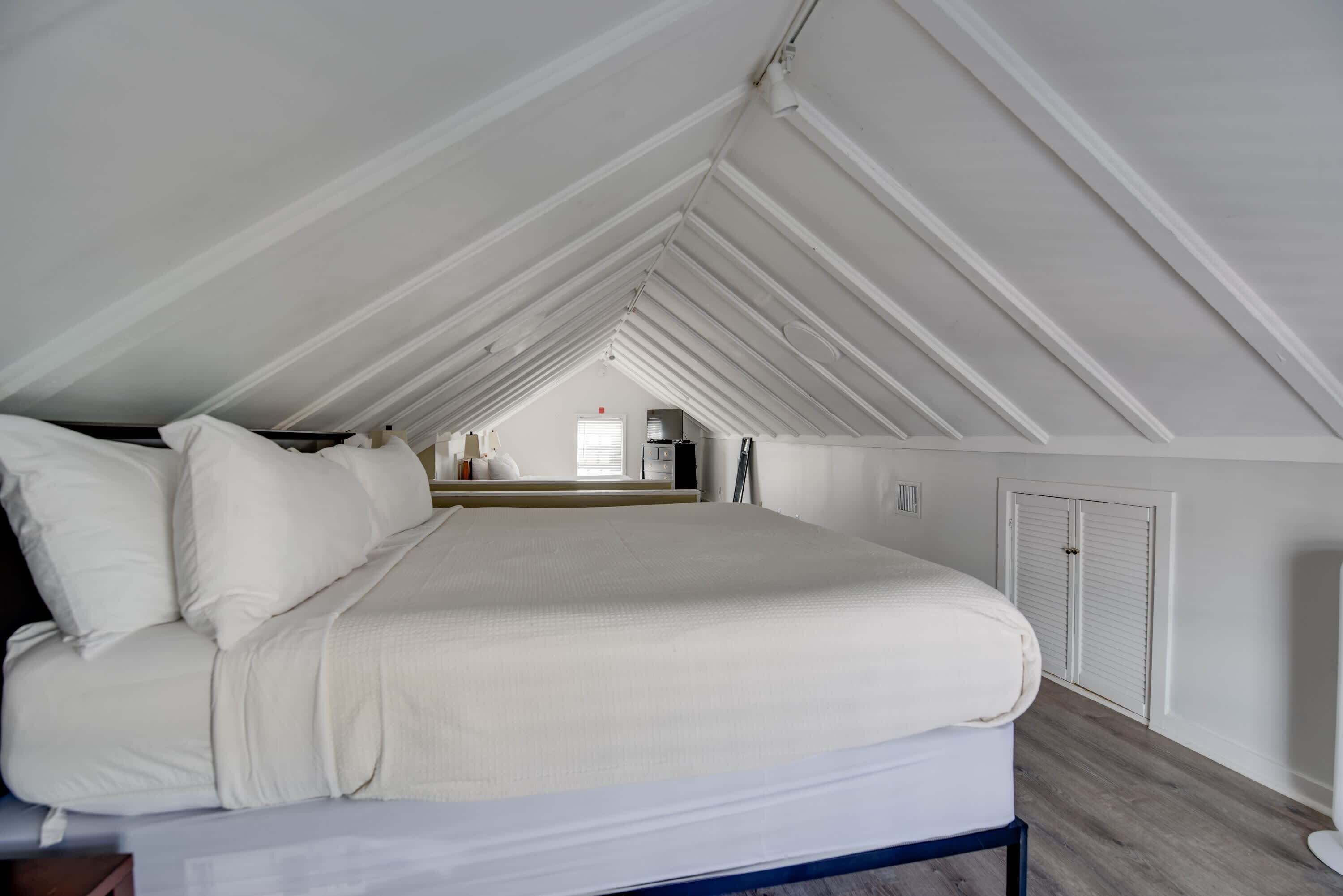 A high-ceilinged attic bedroom with a large bed, white walls, and a view of a kitchen and window.