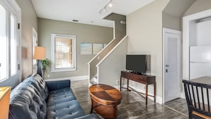 A living room with a leather sofa, a wooden coffee table, a TV on a stand, a staircase, and a door.