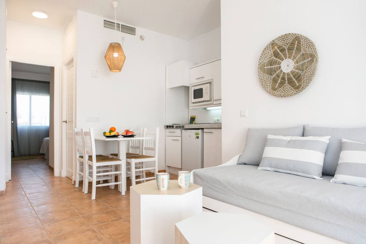 A modern, well-lit living room with a grey sofa, a dining area with a table and chairs, and a kitchen with a microwave.