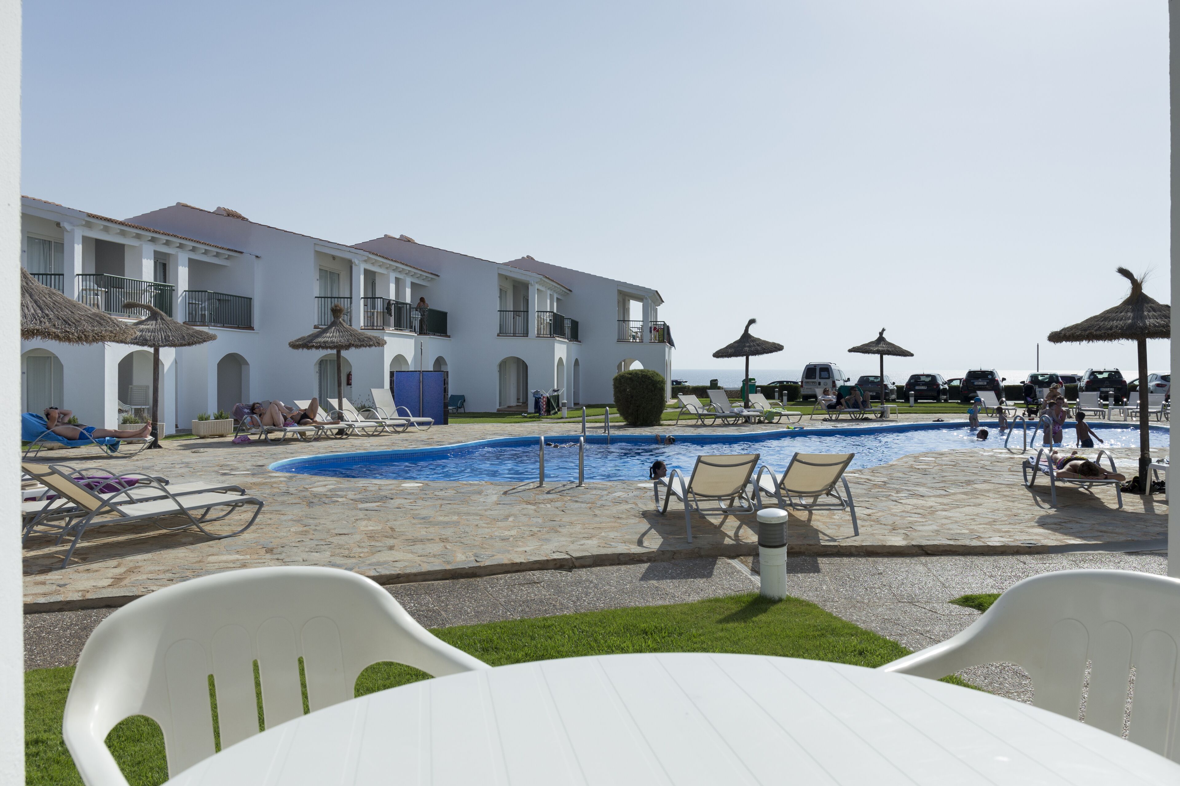 A pool area with lounge chairs and umbrellas, a building with balconies, and a clear sky.