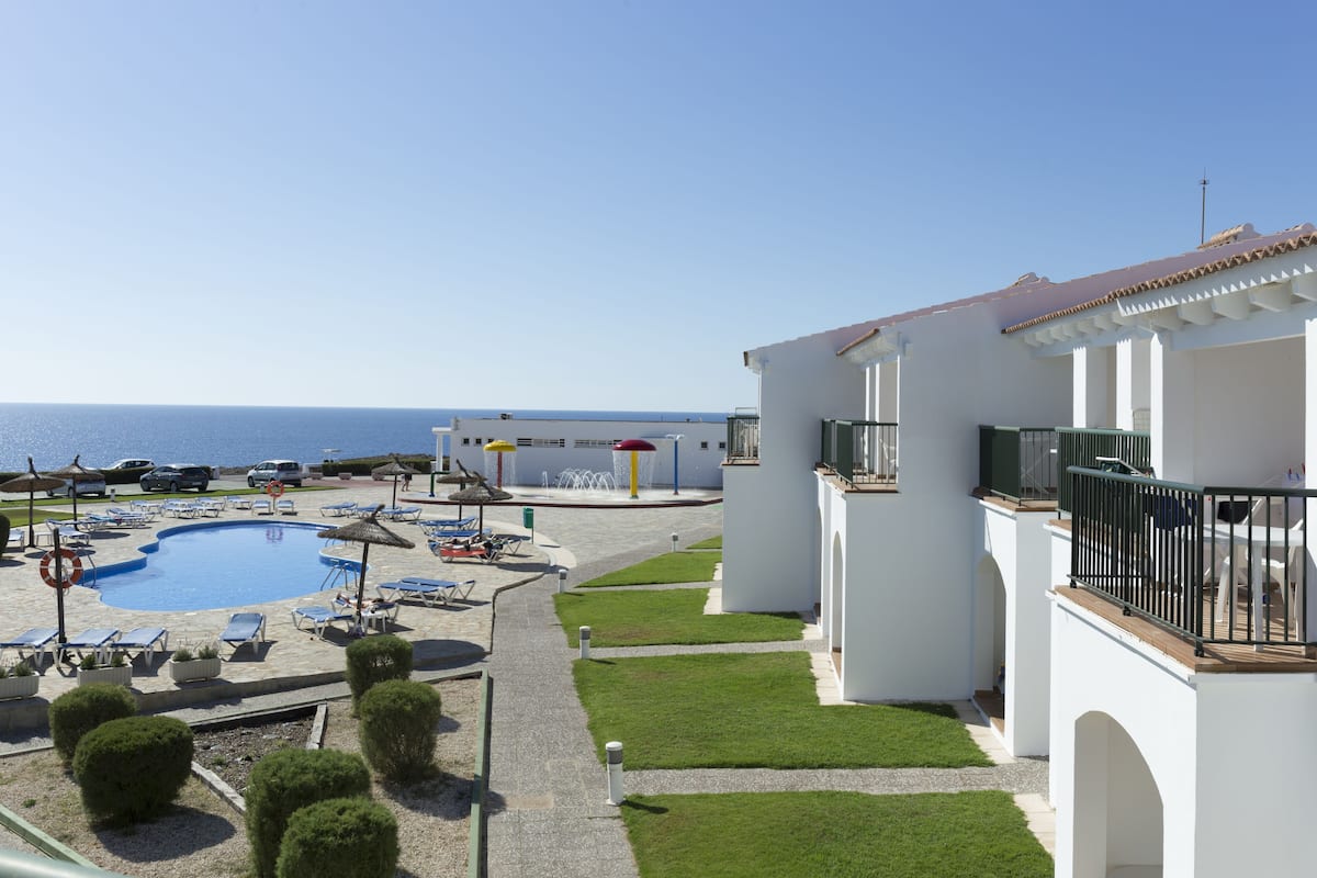 A pool area with lounge chairs and umbrellas, a building with balconies, and a view of the sea.