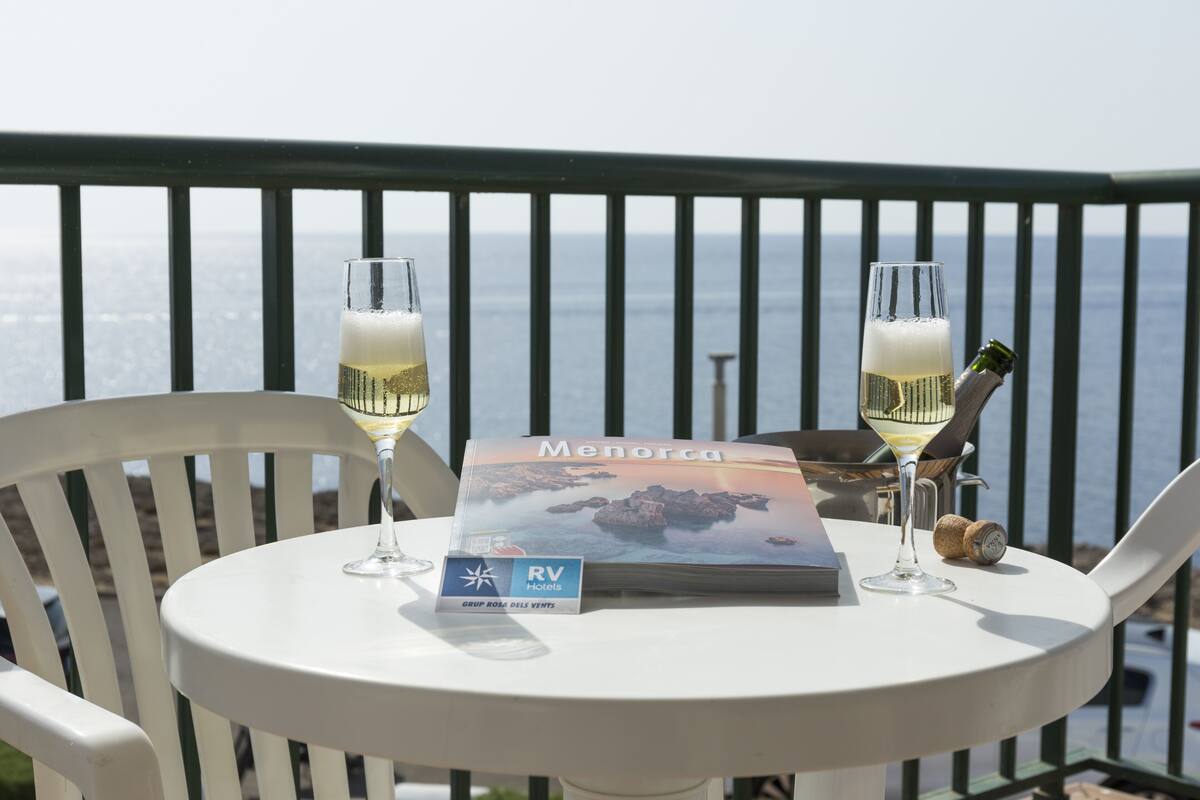 A table with two glasses of white wine, a bottle of champagne, and a brochure on a balcony overlooking the sea.