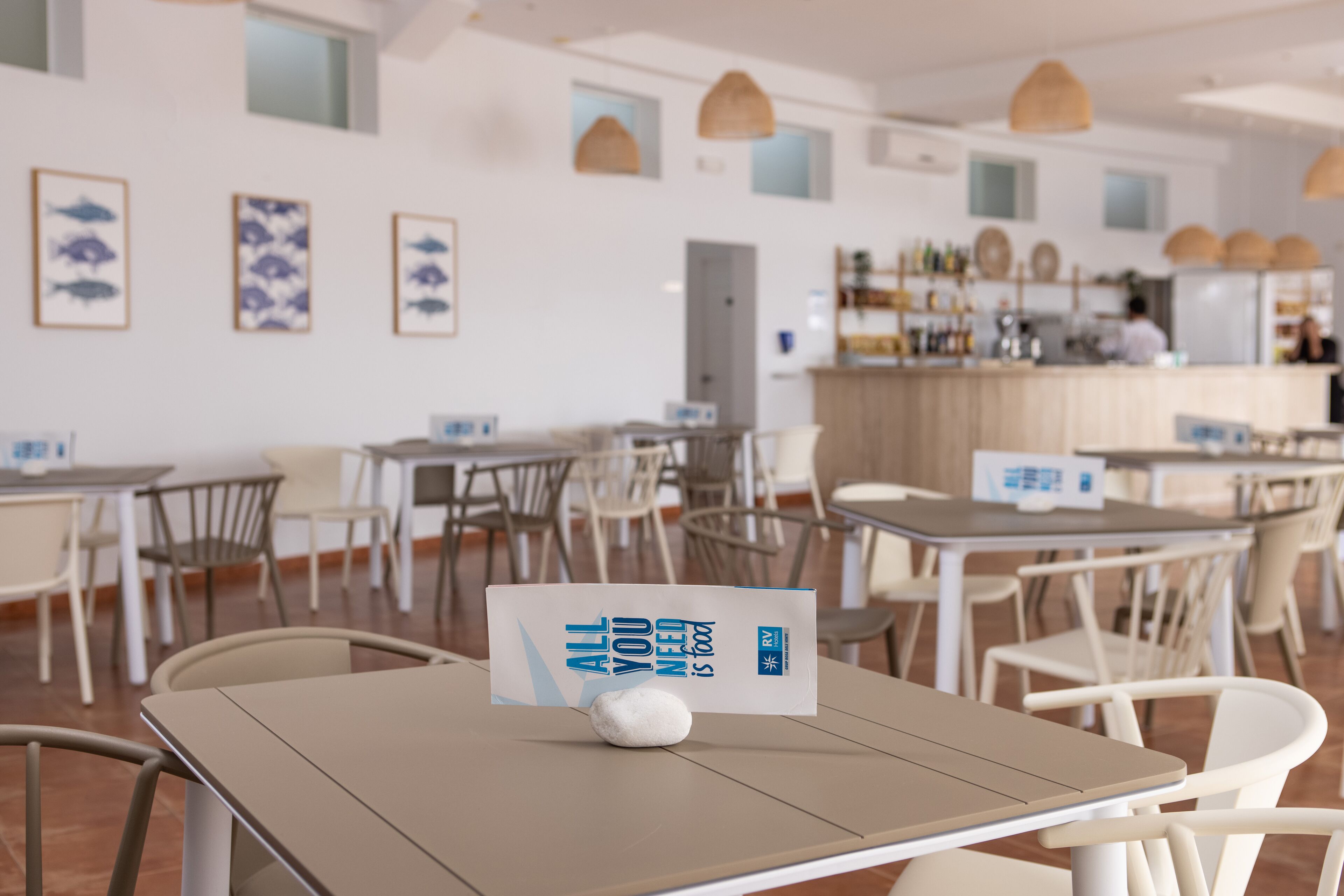 Een modern restaurantinterieur met tafels en stoelen, kunstwerken in lijsten aan de muren en een balie met een persoon achter.