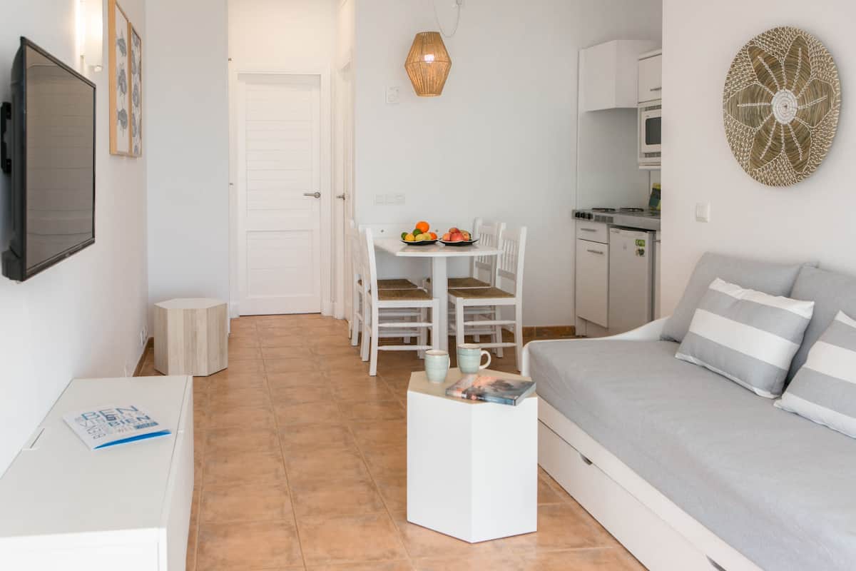 A modern, compact living space with a kitchenette, a dining area with chairs, a sofa with cushions, and a small side table with a book and mug.