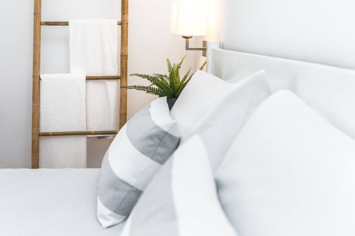 A neatly made bed with white bedding and a grey and white striped pillow. There is a wooden ladder with white towels hanging on it, and a potted plant with green leaves.