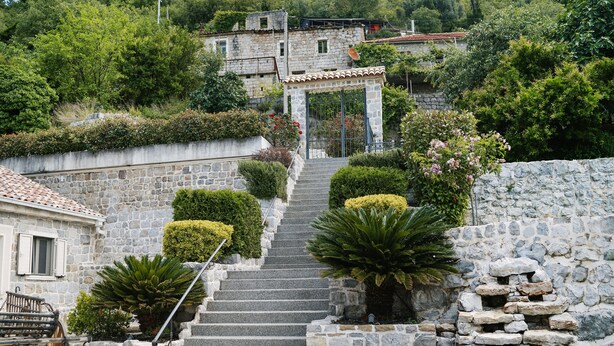 A stone staircase leading to a traditional stone building with a tiled roof, surrounded by lush greenery and a small cart. - Villa Dub (Kotor)