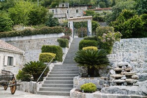 A stone staircase leading to a traditional stone building with a tiled roof, surrounded by lush greenery and a small cart. - Villa Dub (Kotor)