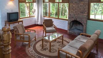 Una sala de estar acogedora con chimenea de ladrillo, asientos confortables, un televisor sobre una mesita y ventanas amplias que dan al exterior.