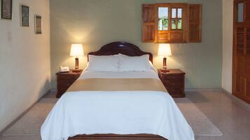 A neatly made bed with a wooden headboard, two bedside tables with lamps, a small window with wooden shutters, and a door with a wooden panel.