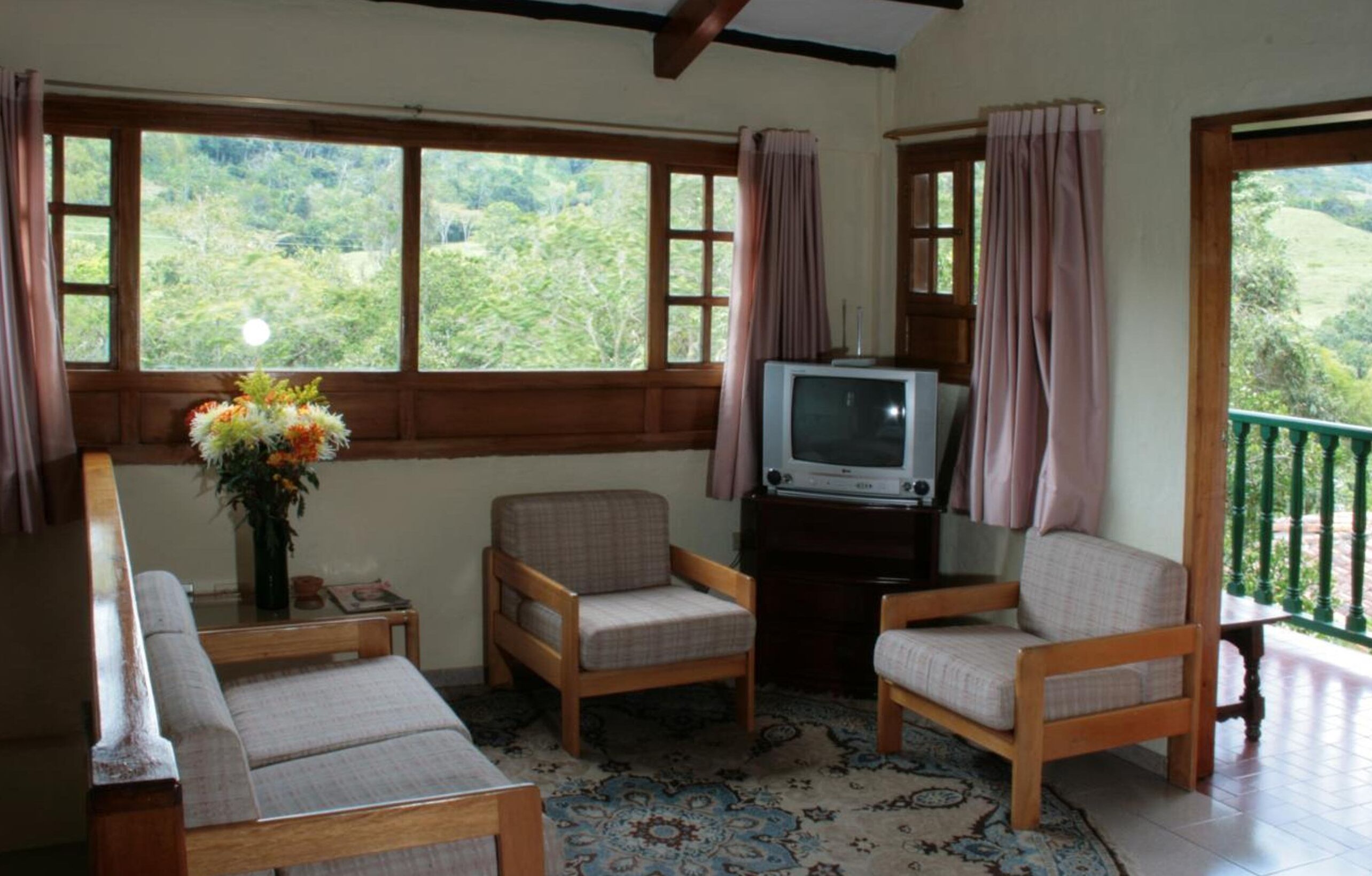 A cozy room with a wooden sofa, two armchairs, a television on a wooden stand, and a large window with curtains.