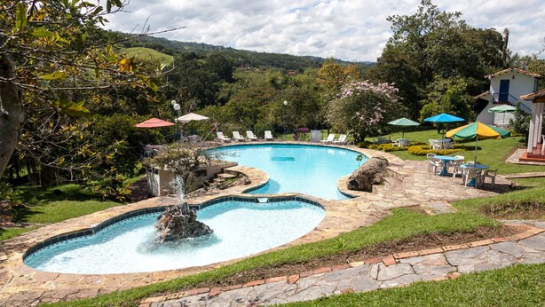 A swimming pool with a waterfall feature, surrounded by lounge chairs and umbrellas, set against a backdrop of lush greenery and hills.