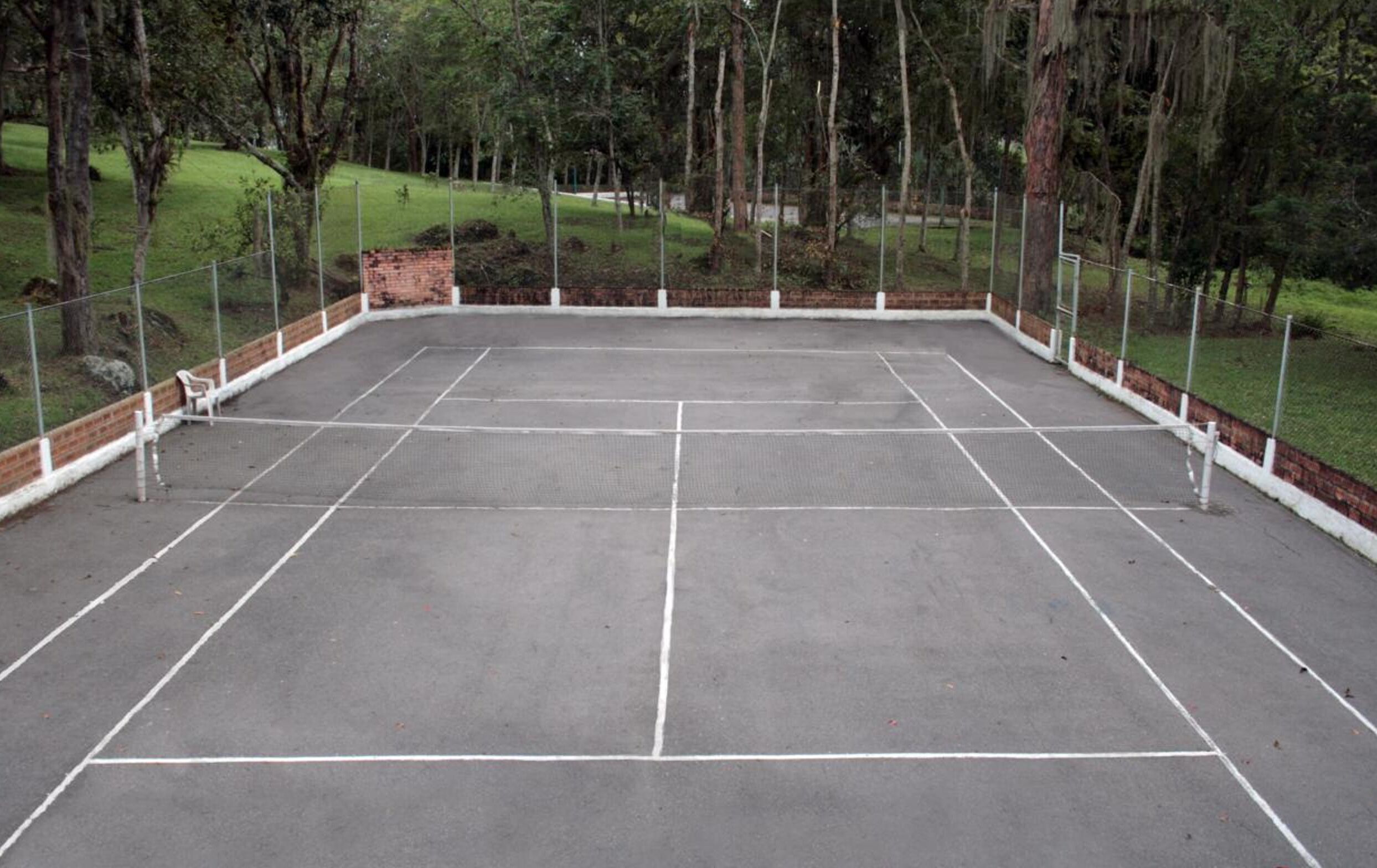 Una cancha de tenis al aire libre rodeada de una valla, con árboles y césped al fondo.