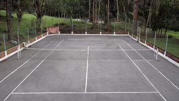 Una cancha de tenis al aire libre rodeada de una valla, con árboles y césped al fondo.