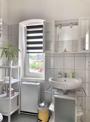 Une salle de bain moderne avec un lavabo blanc, un miroir et un radiateur. Il y a une fenêtre avec des stores et une plante en pot sur une étagère.