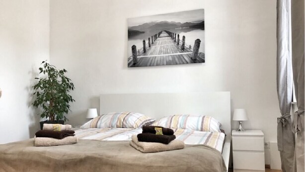 Un dormitorio con cama, una mesita de noche, una lámpara, una planta en maceta y un cuadro en la pared.