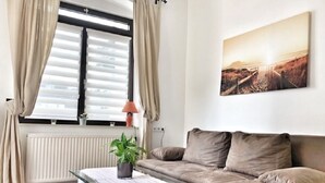 Una sala de estar con un sofá, una mesa con una planta, una ventana con persianas y un cuadro en la pared.
