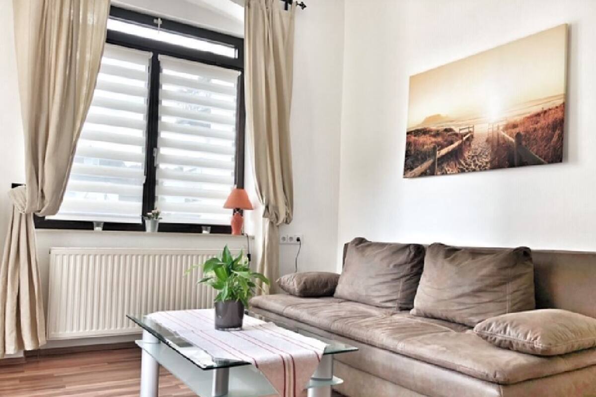 A living room with a sofa, a table with a plant, a window with blinds, and a painting on the wall.