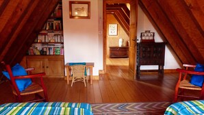 Une chambre confortable située sous les combles, avec des poutres en bois, une bibliothèque, un fauteuil en osier et un lit recouvert d’une literie à rayures.