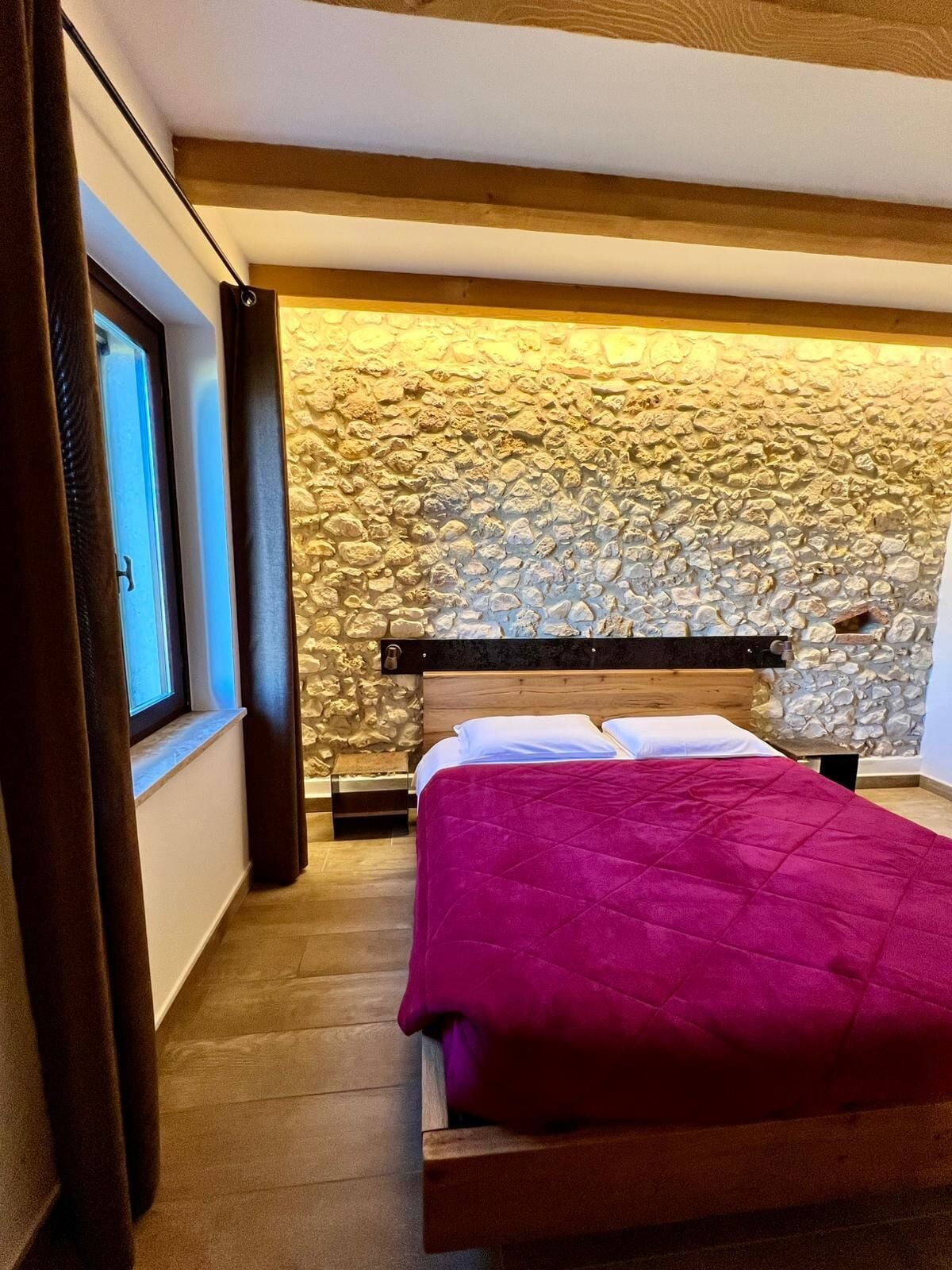 A bedroom with a stone wall, wooden bed frame, and a window with curtains.