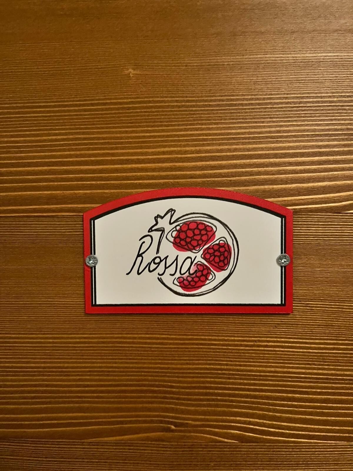 Une surface en bois sur laquelle est apposée une enseigne avec un bord rouge, un fond blanc et une illustration en rouge et noir de framboises, accompagnée du texte « Rosa ».
