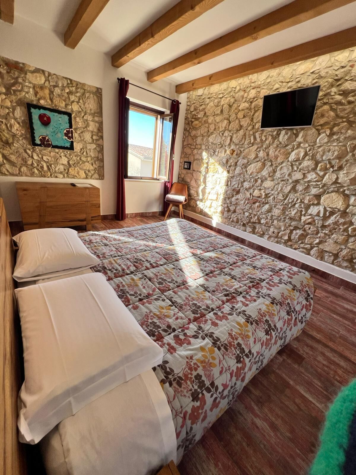 Een slaapkamer met een groot bed, houten balken, stenen muren, een raam met gordijnen en een flatscreen televisie aan de muur.