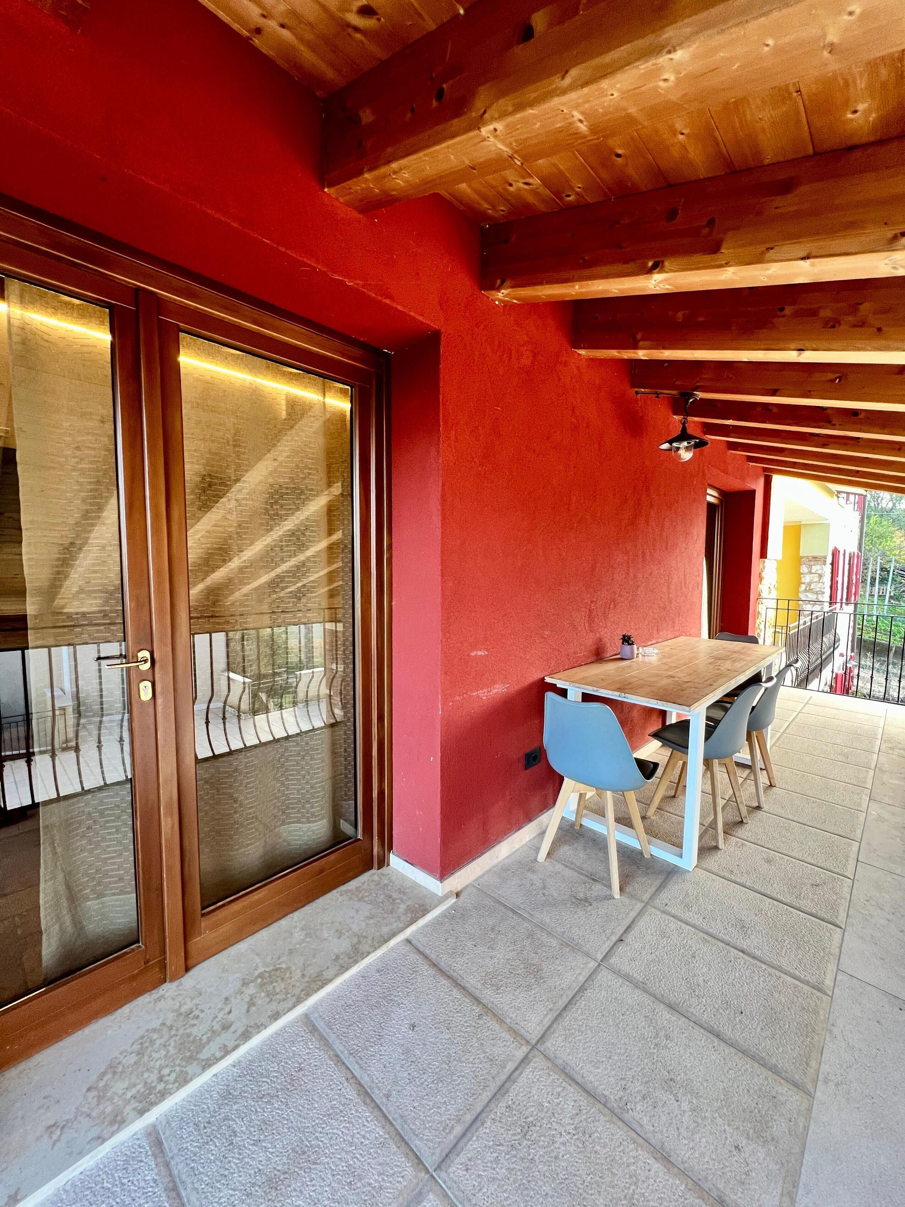 A covered outdoor dining area with a wooden table and blue chairs, a red wall, and a glass door leading to a balcony.