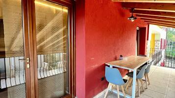 A covered outdoor dining area with a wooden table and blue chairs, a red wall, and a glass door leading to a balcony.