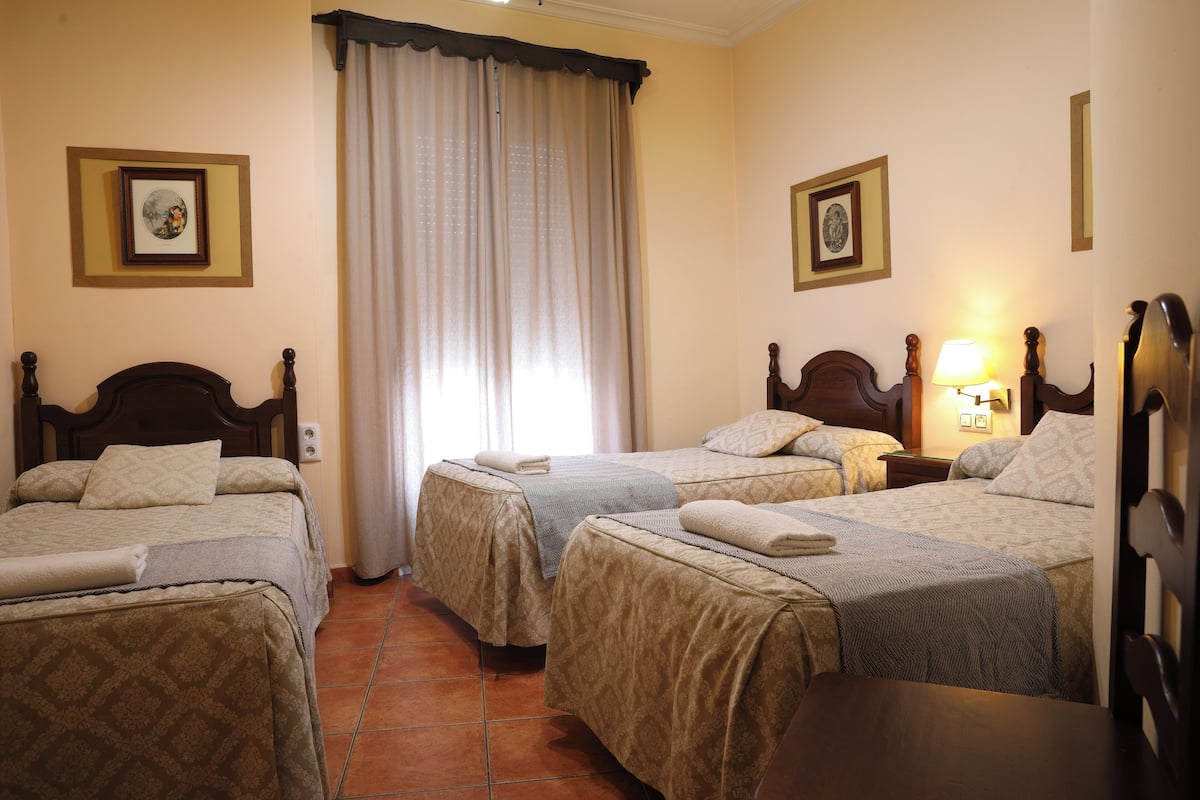 Habitación de hotel con tres camas, un ventanal con cortinas y dos cuadros enmarcados en las paredes.