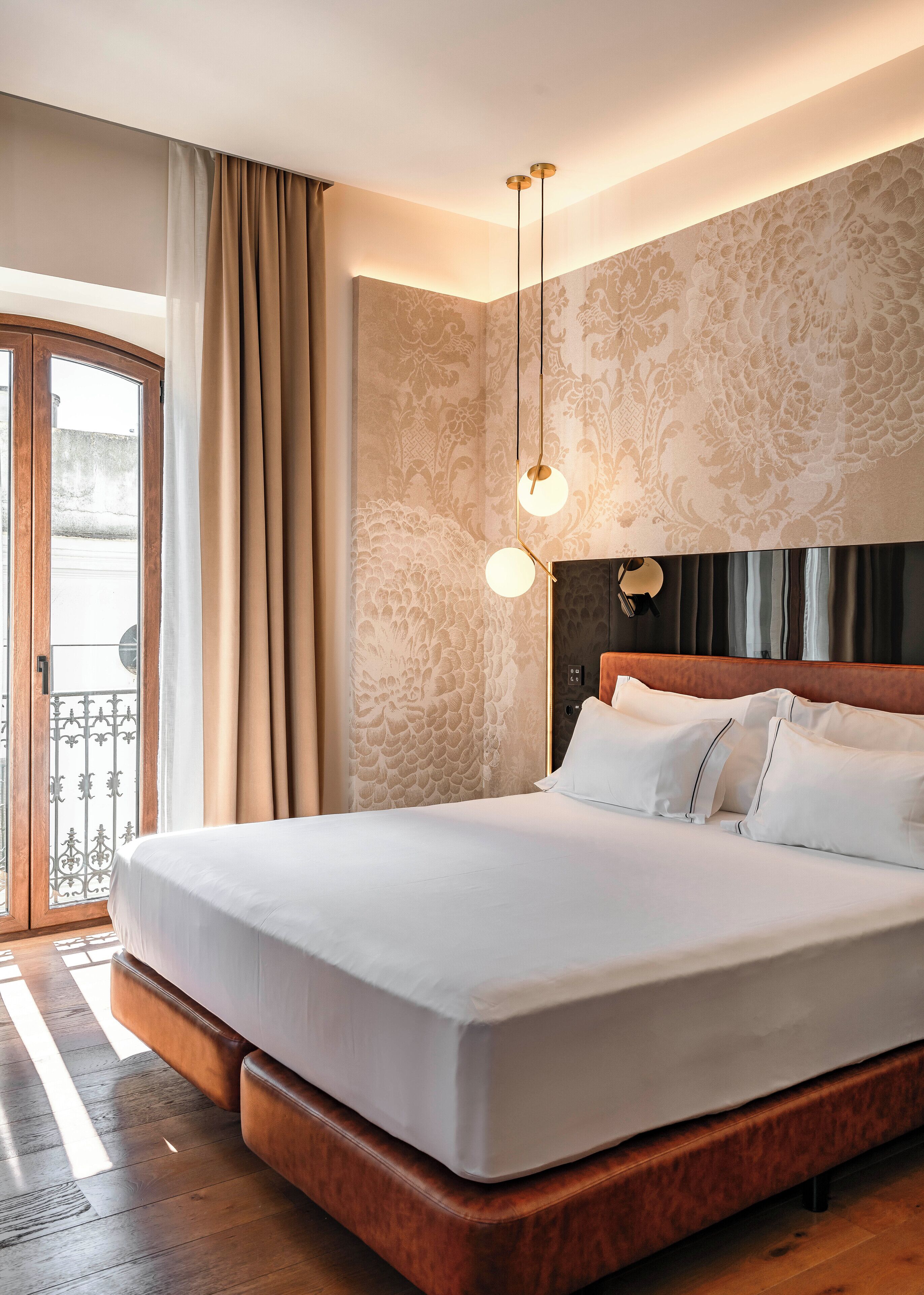 Habitación de hotel con una cama grande, cabecera de madera, pared con estampado y un balcón con cortinas.