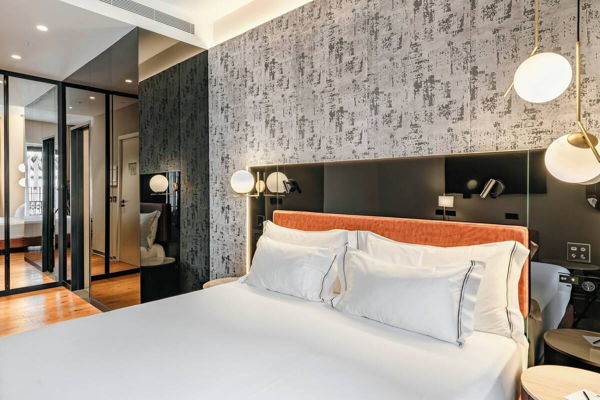 Una habitación de hotel moderna con una cama grande, piso de madera, pared con textura y una mesita de noche con lámpara.