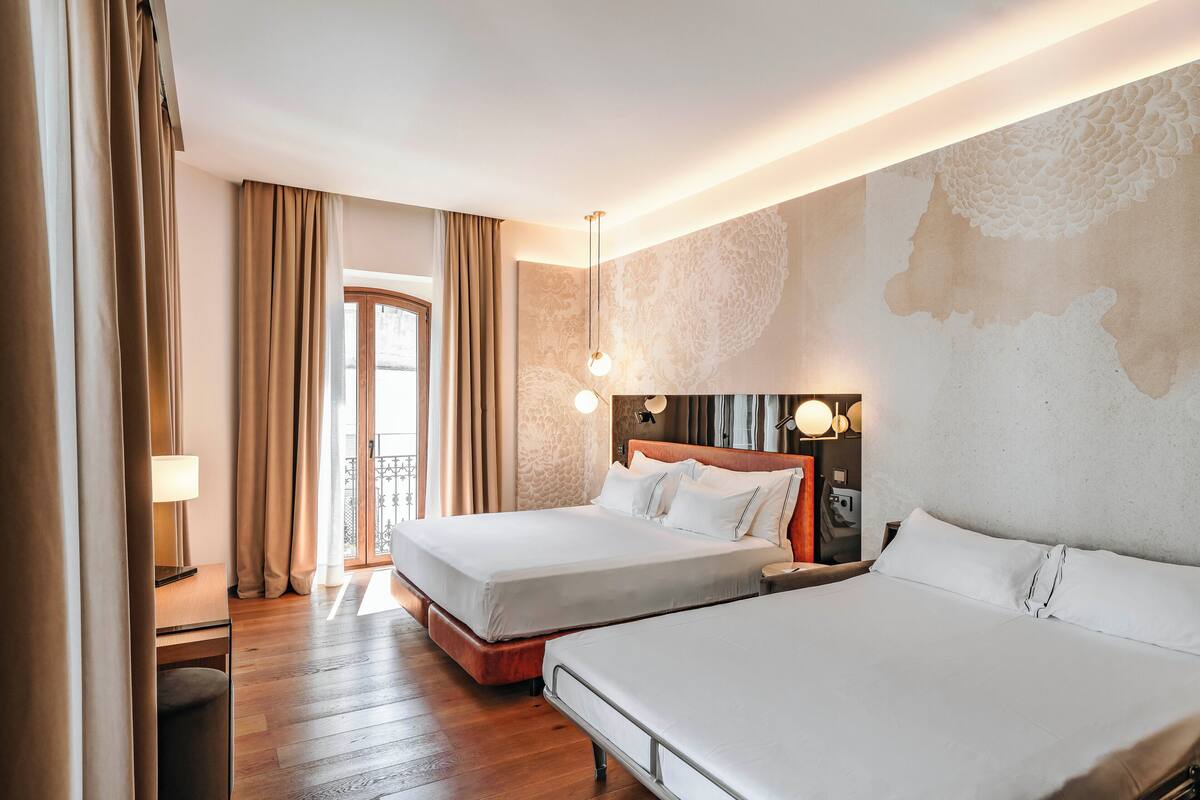 Habitación de hotel con dos camas, piso de madera, un ventanal grande con cortinas y una pared con diseño texturizado.