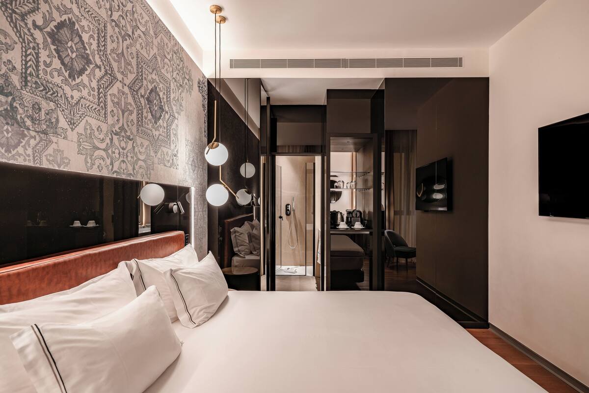 Una habitación de hotel moderna con una cama grande, un vestidor, un baño con espejo y lavamanos, y un televisor instalado en la pared.