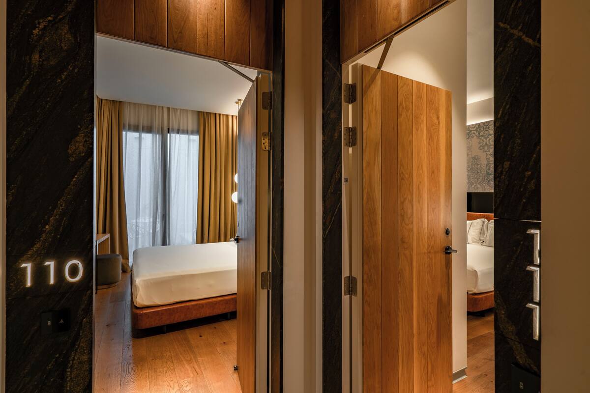 Una habitación de hotel moderna con piso de madera, una cama con ropa blanca, una mesita de noche y un espejo grande. En la pared se observa el número '110'.