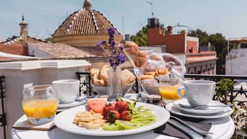 A breakfast table with a variety of food items, including fruits, pastries, and beverages, set on a balcony with a view of a building with a dome and tiled roof.